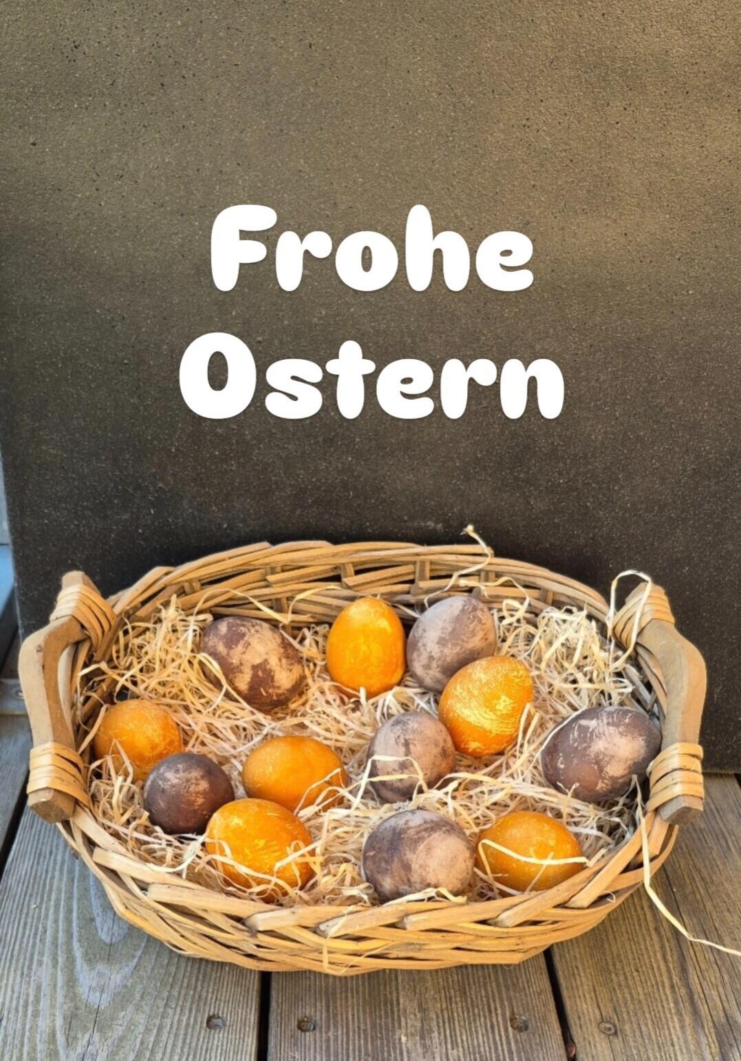 Korb mit bunt gefärbten Ostereiern, umgeben von Osternest-Material, dazu der Schriftzug Frohe Ostern auf Holz.