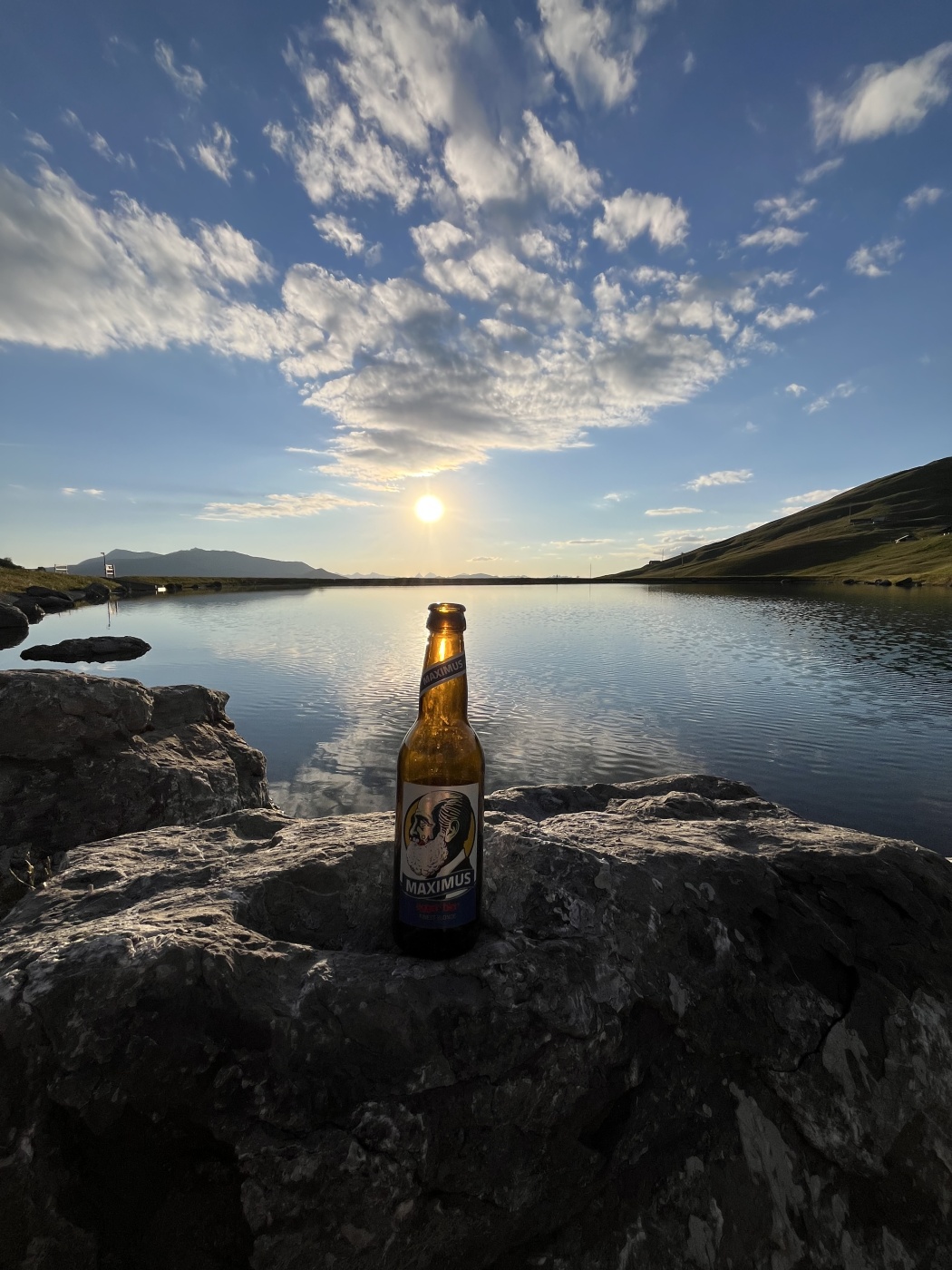 Flasche Bier auf Felsen vor See und Sonnenuntergang, idyllische Naturkulisse mit blauem Himmel und Wolken.